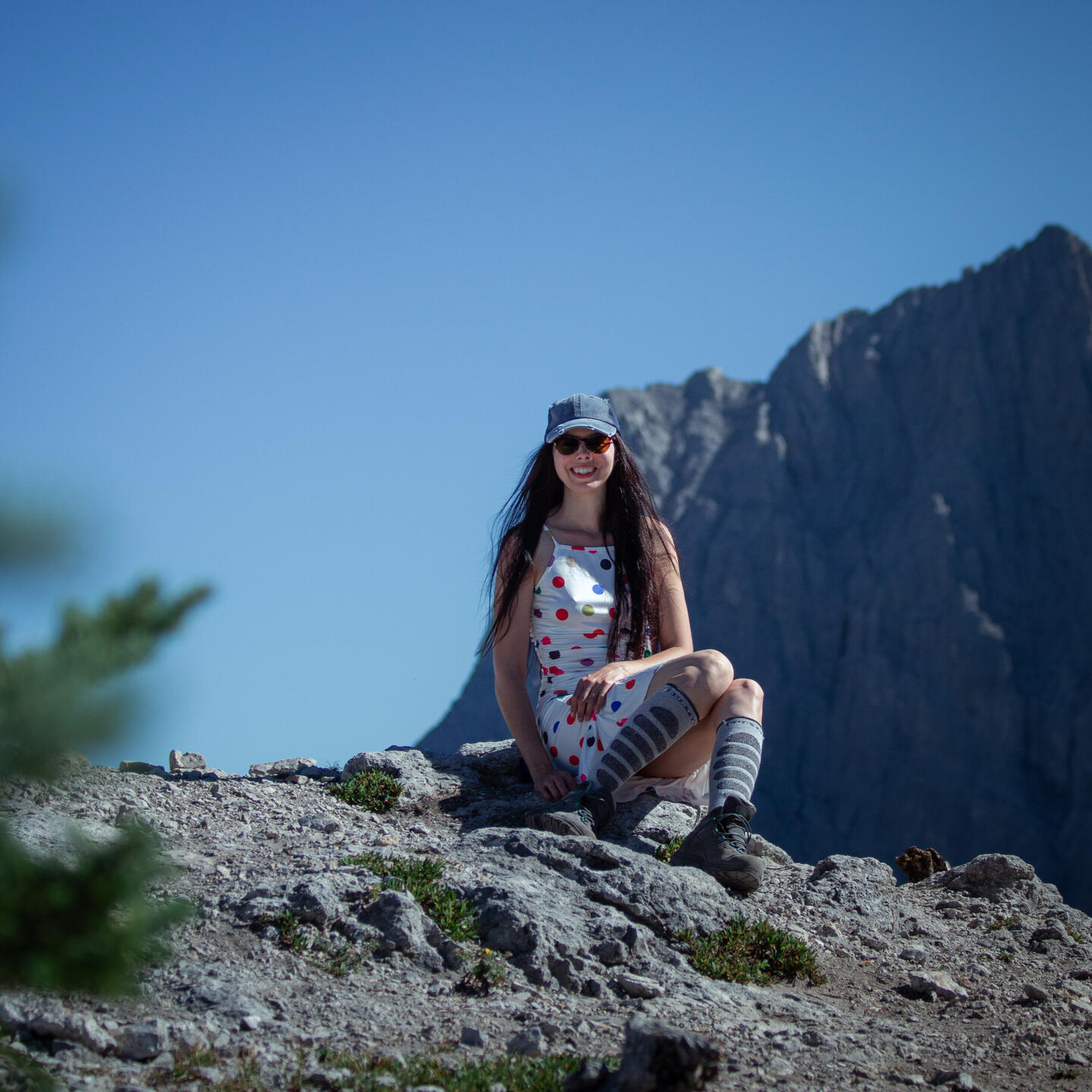 Layla Messner, autistic founder and creator of the Neuro-centered Framework sits on a mountain top. A brunette in a polkadot dress, knee socks, a denim ball cap, and hiking boots sits on a rock in the alpine. Photograph by David Dethlefs.