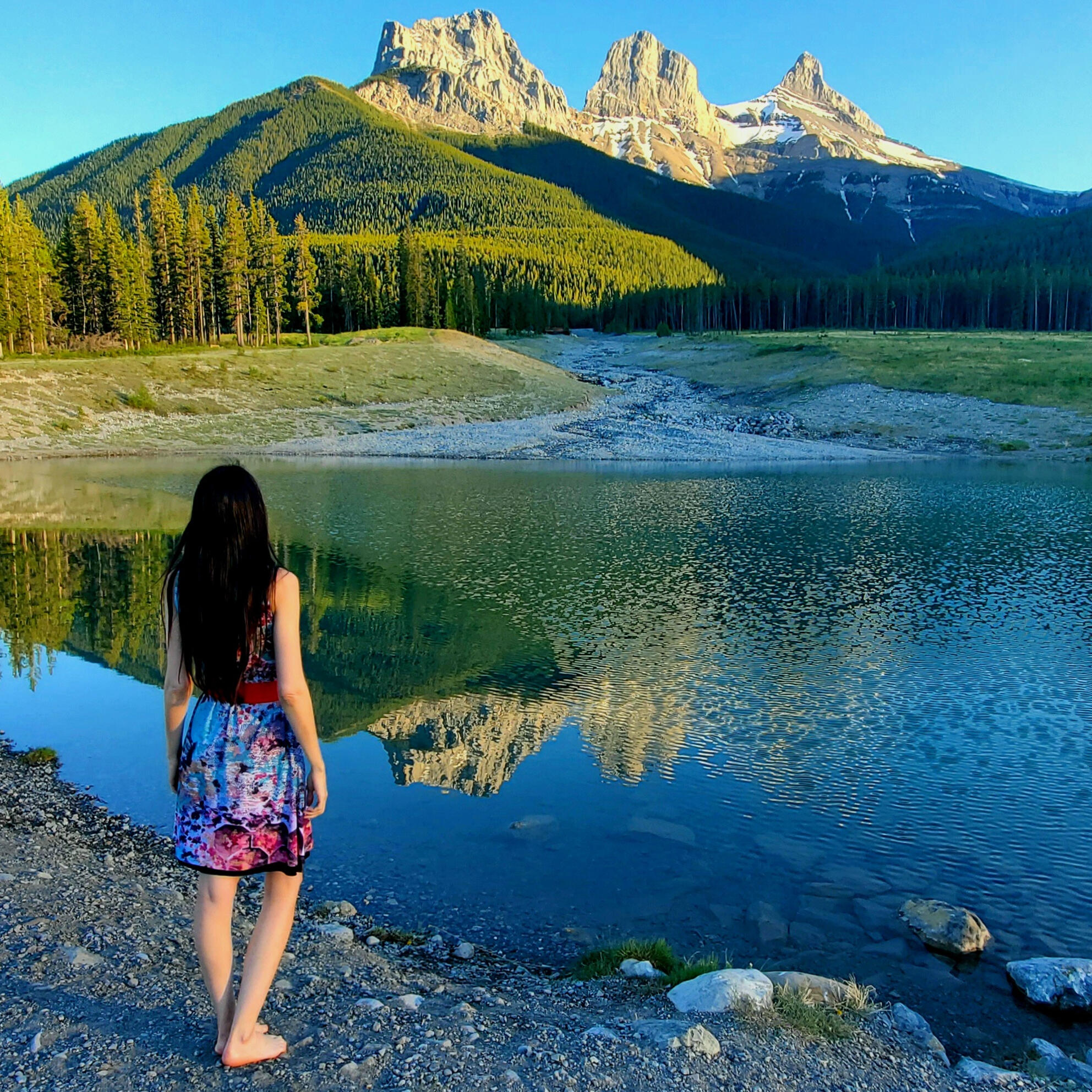 A woman with long dark hair, wearing a colorful dress stands looking at a mountain lake and a clear reflection of the Three Sisters Mountains in Canmore, Alberta, Canada. Photograph by Kelly's Perspective.